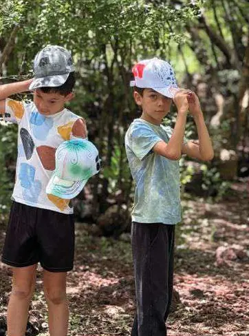 Dos niños con playeras de colores, probándose gorras entre los árboles.