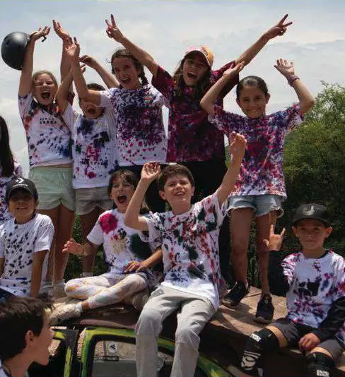 Grupo de niños y niñas con playeras de colores, con los brazos arriba, sobre una camioneta.