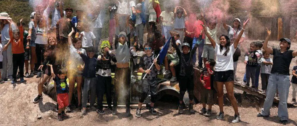 Grupo de niños y adultos con polvos de colores, al aire libre, junto a una camioneta en el parque.
