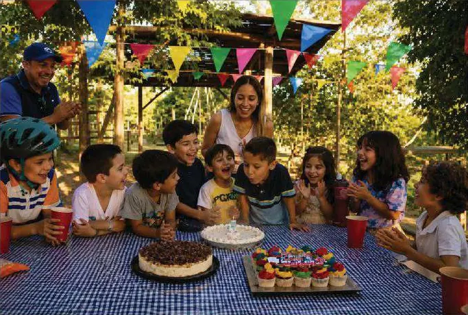 Niños y adultos alrededor de una mesa con pastel, cupcakes y banderines de colores.