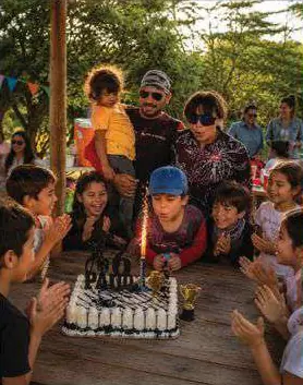 Niños y adultos alrededor de un pastel con una vela, de noche, en una mesa de madera.